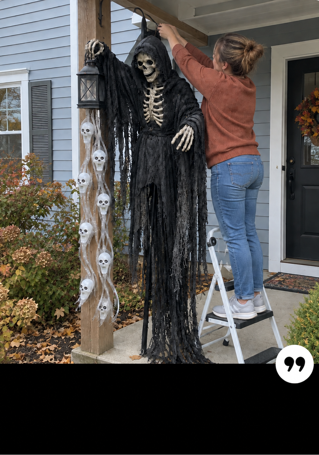 A woman on a white step ladder hangs a large black hooded skeleton Halloween decoration on a porch post outside a house. The decoration has a tattered robe, raised skeletal arms, and visible skull details along the side post.